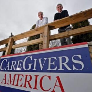 Two people standing behind a 'CareGivers America' sign outdoors.