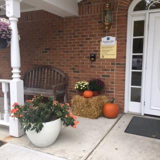 A cozy porch decorated for autumn with pumpkins, a hay bale, and colorful flowers.