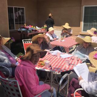 A group of people wearing wide-brimmed hats sit around tables with checkered tablecloths in an outdoor setting.