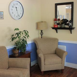 A cozy living room corner featuring two armchairs, a decorative lamp, a clock on the wall, and a plant.