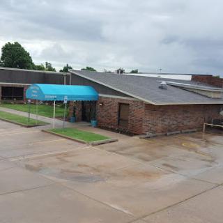 A view of a brick building with a blue canopy, surrounded by a parking area under a cloudy sky.