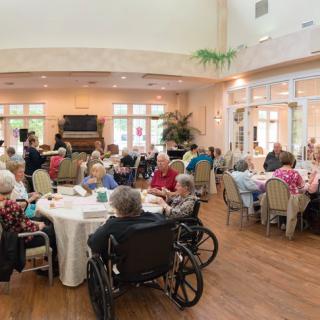 A group of seniors gathered in a dining hall, some using wheelchairs, enjoying a communal event.