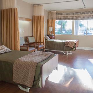 A hospital room with two beds, chairs, and large windows.