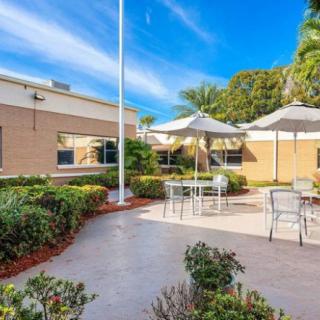 A sunny outdoor patio area with tables, chairs, and umbrellas surrounded by a landscaped garden.