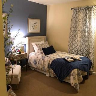 A cozy bedroom with a single bed, blue and floral bedding, a nightstand with a lamp, and patterned curtains.