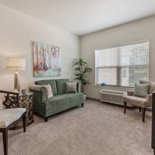 A cozy living room with modern decor, featuring a green sofa, chairs, floor lamp, artwork, and large window.