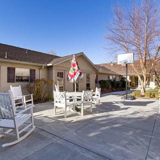 A sunny patio with rocking chairs, a table with an umbrella, and a basketball hoop in front of a house.
