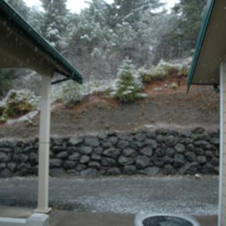 A snowy landscape with trees and a rock wall between two structures.