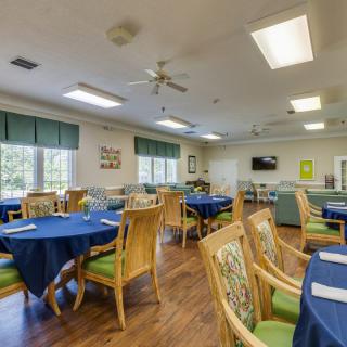 A spacious dining room with blue tablecloths, wooden chairs, and ample natural lighting from large windows.