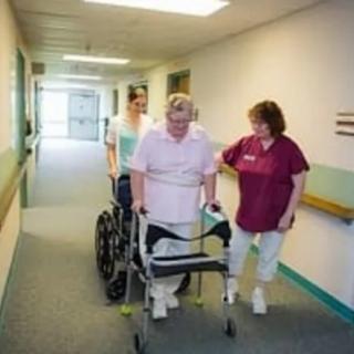 Healthcare professionals assist an elderly patient with mobility in a nursing home hallway.