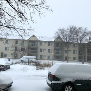 A snowy scene featuring an apartment building surrounded by snow-covered cars.