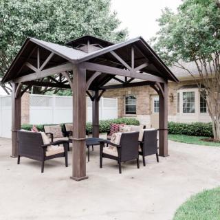 A cozy outdoor patio area featuring a wooden gazebo with comfortable wicker chairs and a table, surrounded by lush greenery.