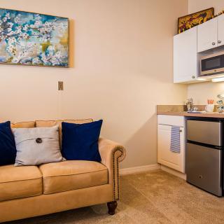 A cozy living area featuring a beige sofa with blue pillows next to a kitchenette, adorned with a piece of abstract artwork.