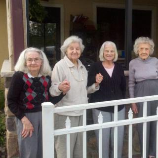 A group of elderly women standing together on a porch behind a white fence.