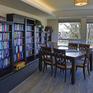 A cozy reading room with bookshelves, a table, and chairs by large windows.