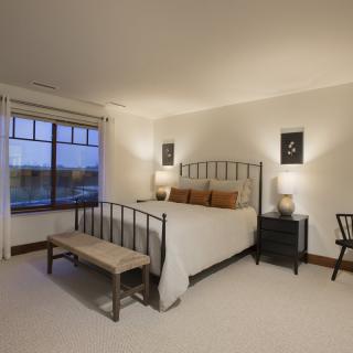 A cozy bedroom featuring a metal-framed bed, two side tables with lamps, a bench at the foot of the bed, and a large window.