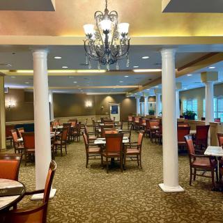 An elegant dining room with arranged tables and chairs, featuring a chandelier and decorative pillars.