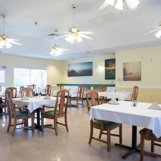 A bright and spacious dining room with neatly arranged tables covered in white tablecloths, complemented by wooden chairs and ceiling fans overhead.