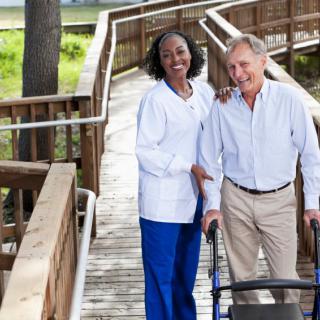 A nurse assists an elderly man with a walker on a wooden pathway outdoors.