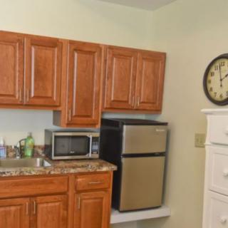 A compact kitchen area with wooden cabinets, a microwave, mini fridge, sink, and a wall clock.