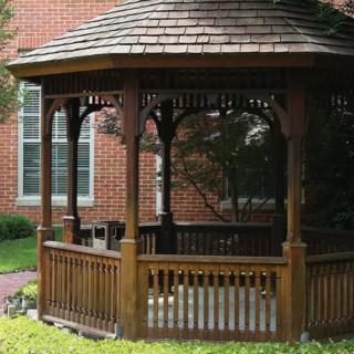 A wooden gazebo situated in front of a brick building within a garden setting.