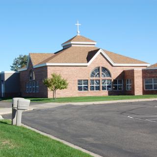 A brick church building with a cross on the roof, large windows, and a parking lot in front.