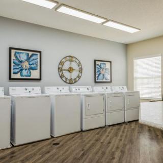 A bright laundry room featuring multiple washing machines and dryers with decorative artwork and a wall clock.