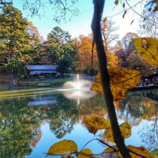 A tranquil autumn scene featuring a pond with a fountain, surrounded by colorful foliage.