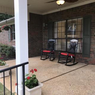 A cozy porch with two black rocking chairs and red cushions, surrounded by potted plants and a brick exterior.