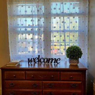 A wooden dresser topped with a 'welcome' sign and an indoor plant, situated in front of a window with lace curtains.