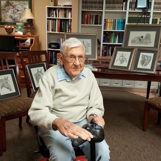 An elderly man sits in a library with framed art pieces on display behind him.