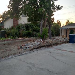 A residential yard with a pile of rocks along the driveway beside trees and trash bins.