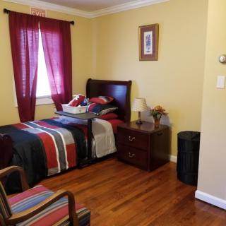 A cozy bedroom with a single bed featuring colorful striped bedding, a wooden nightstand with a lamp, and rich wood flooring. The window is adorned with red curtains.