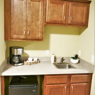 A compact kitchenette with wooden cabinets, a countertop featuring a coffee maker, sink, and a mini fridge below.