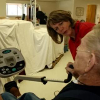 A healthcare professional assists an elderly person using exercise equipment during a physical therapy session.