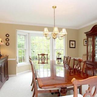 A traditional dining room with a wooden table, elegant chairs, and a chandelier under natural light from a bay window.