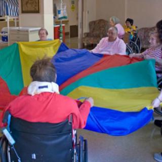 A group of seniors in wheelchairs participate in a colorful parachute activity indoors.