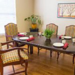 A cozy dining room featuring a wooden table with chairs, adorned with plates and red napkins, complete with a potted plant centerpiece and wall artwork.