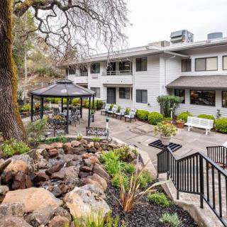 A serene outdoor garden with a gazebo and seating area, surrounded by lush greenery and a rock feature in front of a two-story building.