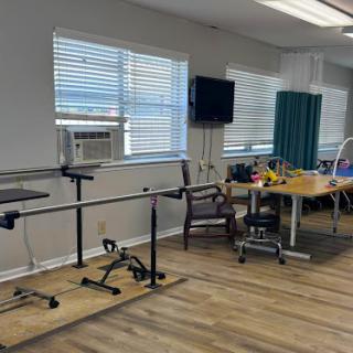 A spacious physical therapy room equipped with a railing for walking assistance and therapy tables, featuring natural light from large windows.