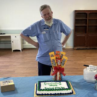 A man in a blue shirt stands beside a decorated birthday cake, celebrating a special occasion.