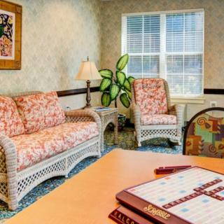 A cozy living room featuring a wicker sofa and armchair with patterned cushions, a small table with a lamp, an indoor plant near the window, and a game board on the table.
