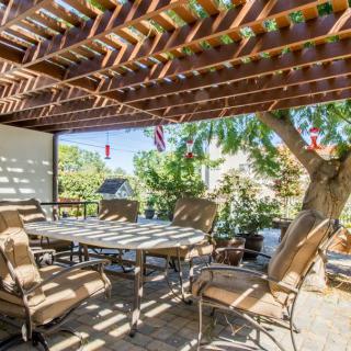 A cozy outdoor dining area under a wooden pergola with comfortable seating and surrounded by greenery.
