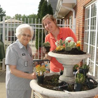 Two women gardening together, planting flowers in a large outdoor planter.