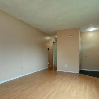 An empty room with wooden flooring and neutral-colored walls, leading to a hallway.