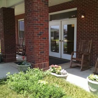 A welcoming entrance to a brick building, featuring glass doors, rocking chairs, and vibrant planters with flowers.