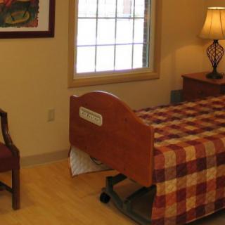 A well-lit room with a hospital bed covered in a checkered quilt, alongside a chair and a lamp.