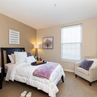 A cozy bedroom with a neatly made bed, a bedside lamp, and an armchair near the window.
