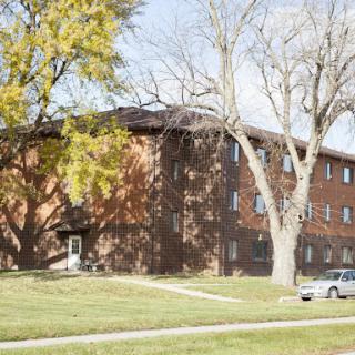 A brick apartment building surrounded by trees and grass, featuring a parked car in the foreground.