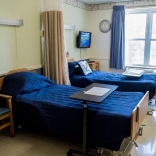 A hospital room with two patient beds, a curtain for privacy, a television on the wall, and a window providing natural light.